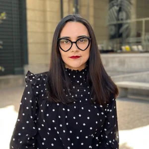 Retrato de Cynthia Herrera, una mujer con lentes de estilo cat-eye, blusa negra de lunares blancos y labial rojo.
