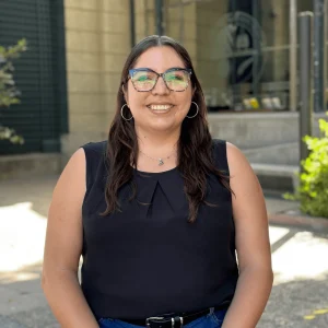 Retrato de Maria Ignacia Salas, una mujer joven con lentes, blusa negra y jeans, sonriendo al aire libre.