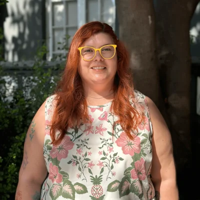 Retrato de Stefani Mardones, una mujer de cabello pelirrojo con lentes amarillos y blusa con estampado floral, sonriendo al aire libre.